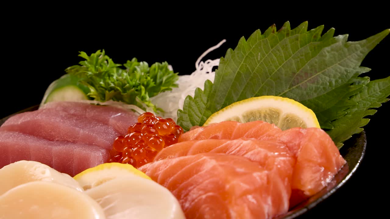 A vibrant assortment of salmon roe, scallop, tuna, and salmon sashimi arranged with garnish on a plate, smoothly rotating against a black background under studio lighting