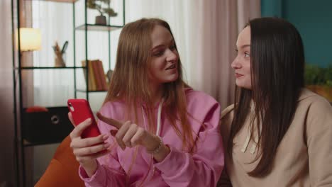 cheerful girls friends siblings watching photos on smartphone on sofa at home, enjoy social media