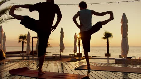 two guys do yoga and stand on one leg on the beach in the morning. silhouettes of people at sunrise