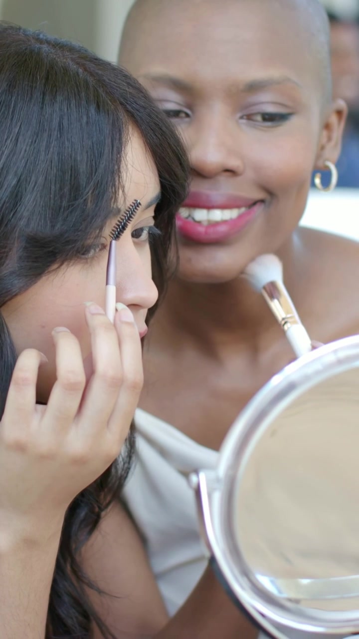 Vertical video: Leaning two women doing brows blending cheeks at mirror with spoolie brush prepping
