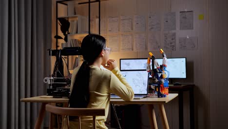 back view of teen asian girl with 3d printing thinking while designing a cyborg hand on a laptop at home