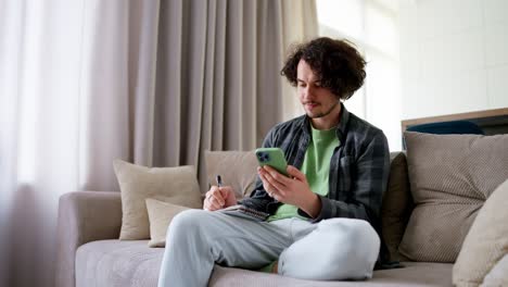 A-confident-brunette-guy-with-curly-hair-in-a-gray-checkered-shirt-takes-notes-and-notes-that-he-watches-a-video-on-a-green-smartphone-at-home-on-the-sofa-in-a-modern-apartment