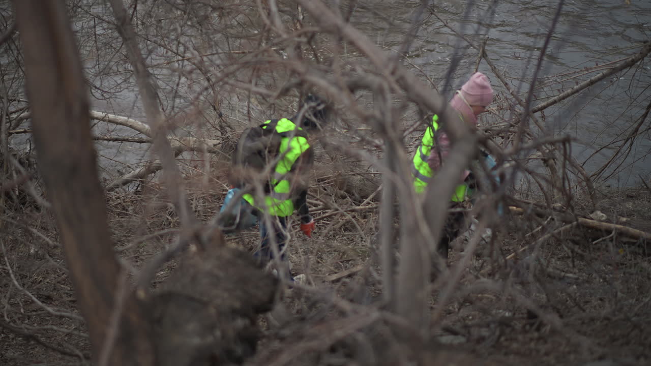Volunteers wearing bright reflective vests collecting trash along polluted riverbank, removing plastic bottles and litter from muddy ground among bare trees during cold season to protect environment