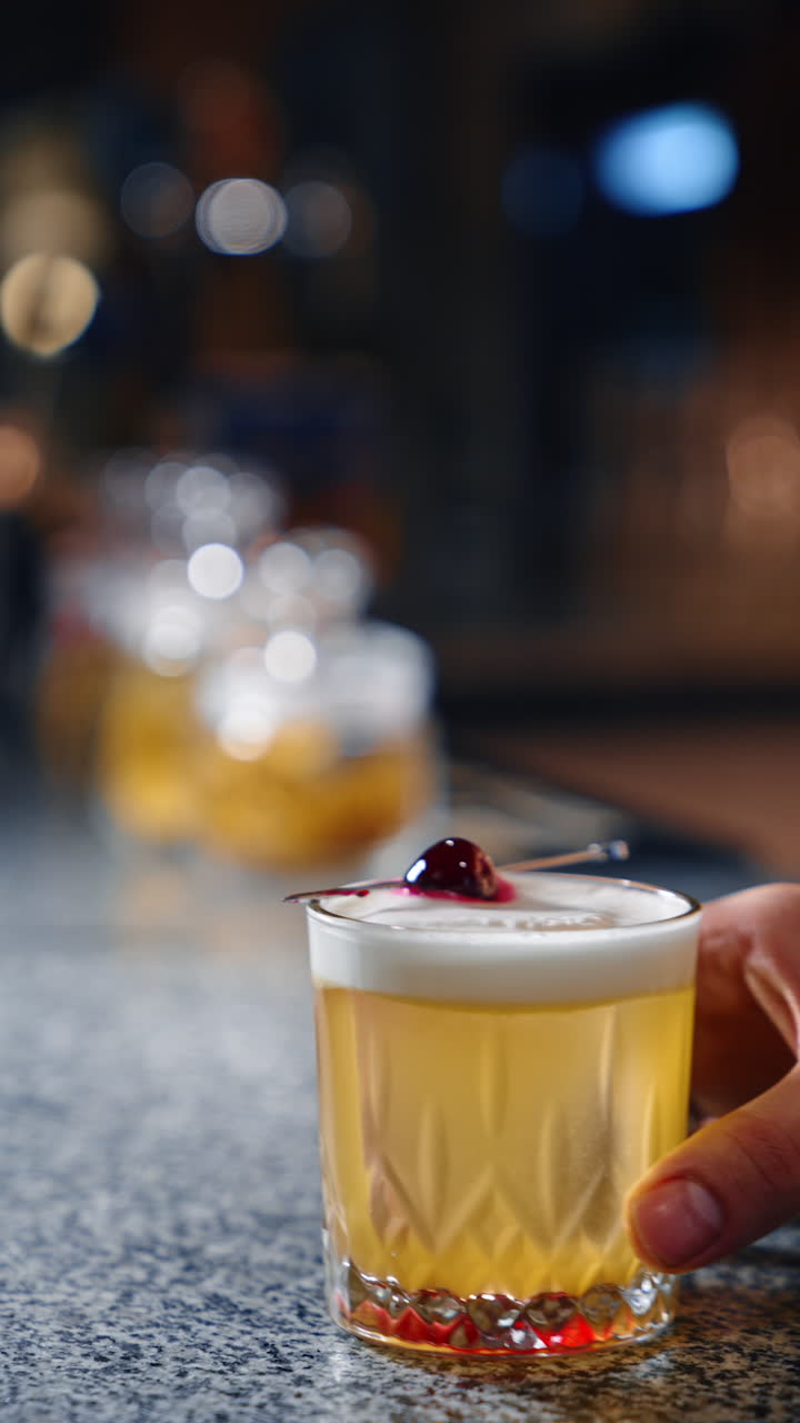 Male hand pushes the glass with a yellow drink by the bar stand. A toothpick with cherry on top. Close up. Vertical video.