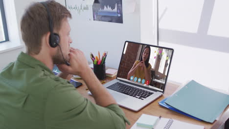 ein kaukasischer mann mit telefonkopfhörer hat einen videoanruf mit einer weiblichen kollegin auf dem laptop im büro