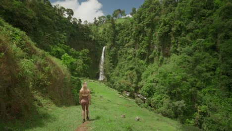 Mujer-En-Ropa-Deportiva-Caminando-Cuesta-Abajo-Hacia-Una-Cascada-Tropical-En-Un-Exuberante-Valle-Verde