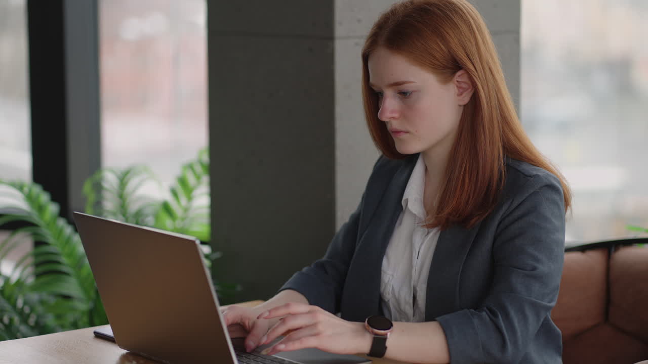A young red-haired woman is working on a laptop. Remote work. Home office. Office work. A woman in a suit in the office is typing on a laptop