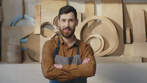 portrait of caucasian bearded man in apron smiling at camera with arms crossed in carpentry workshop