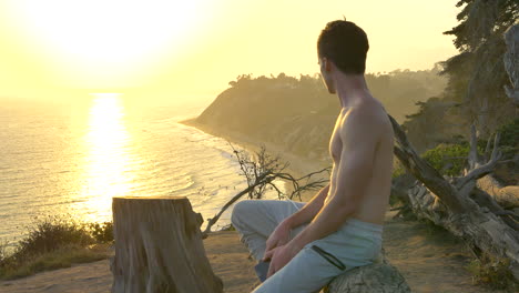 joven atractivo hombre sin camisa descansando después de un entrenamiento físico en el borde de un acantilado sobre el océano pacífico durante una puesta de sol dorada en santa barbara, california