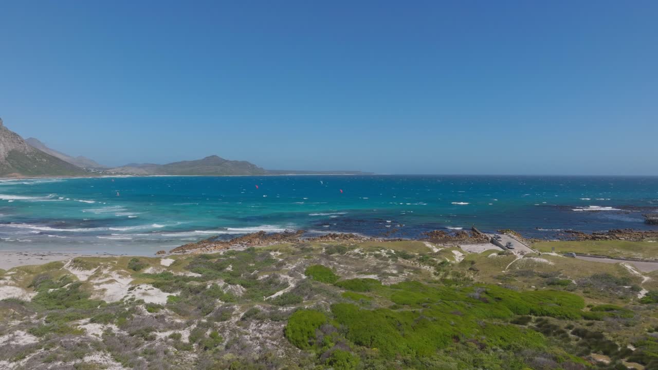 Drone glides towards the rugged shoreline of Misty Cliffs village near Cape Town, revealing deep blue Atlantic Ocean waters, sandy beaches, and coastal vegetation under a clear sky