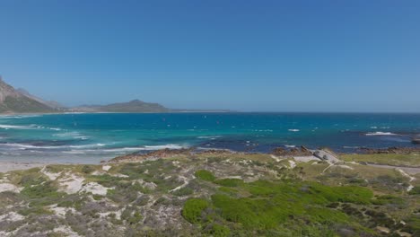 drone glides towards the rugged shoreline of misty cliffs village near cape town, revealing deep blue atlantic ocean waters, sandy beaches, and coastal vegetation under a clear sky