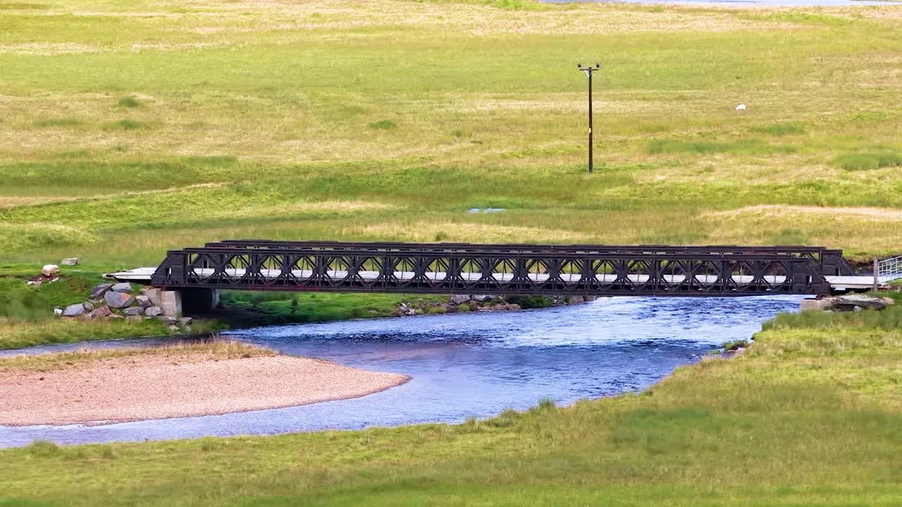 Dronebeelden tonen een donkere metalen brug over een blauwe rivier in een weelderige vallei
