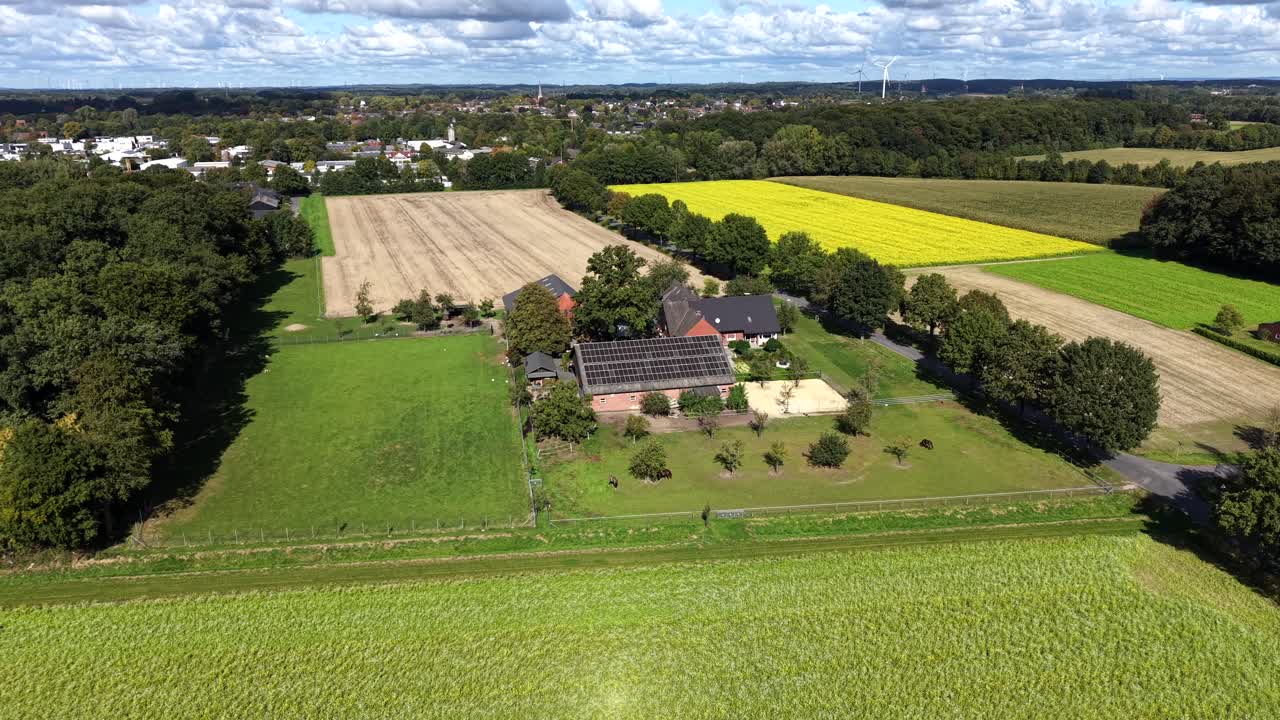 Aerial flyover German farmstead with barn and solar panels on roof. Rural suburb district. Wheat field, crop and corns. Yellow rape field and maize in late summer season. Tree-lined road. Wide shot