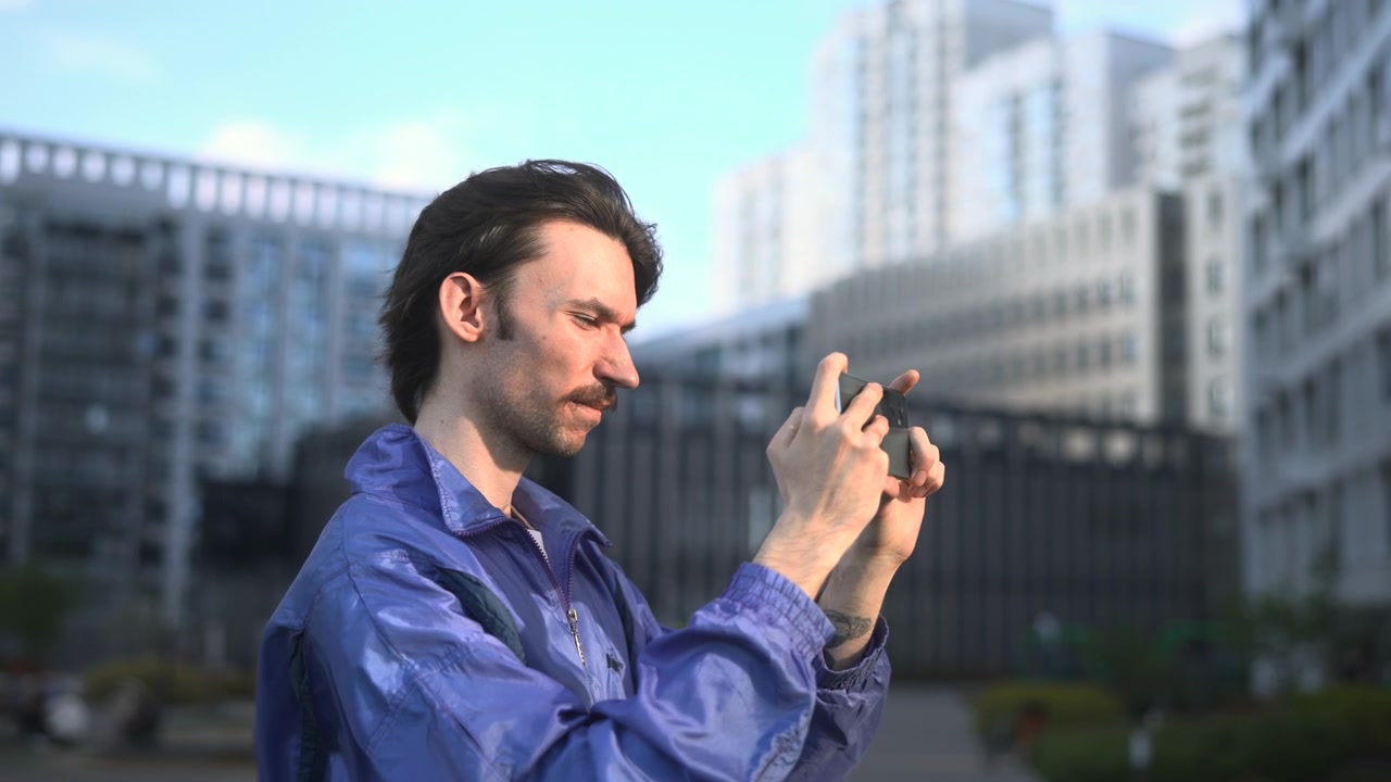 Giovane uomo con una giacca blu sta in piedi in primo piano usando uno smartphone, con edifici urbani sfocati sullo sfondo