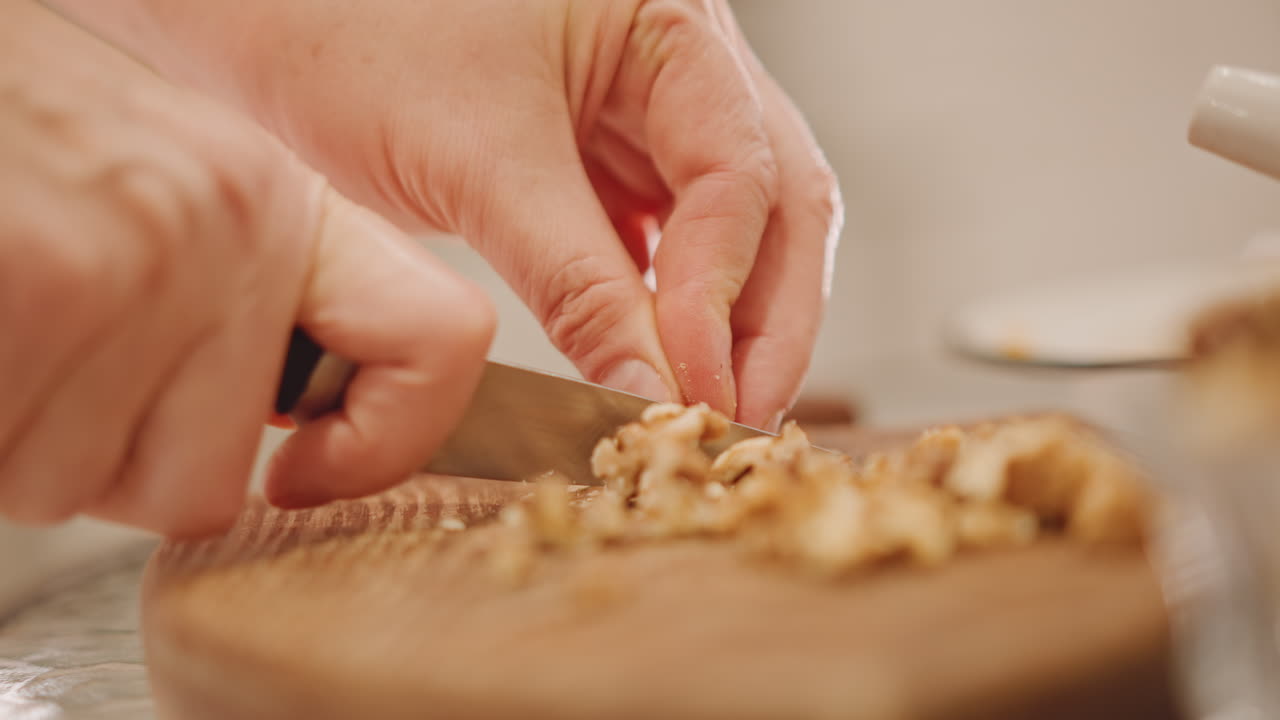Closeup hands chopping walnuts on wooden board, precise knife cuts, fine mince for pumpkin pie filling, textured crumbs, slow deliberate motion, skilled home baker prep, rustic countertop light