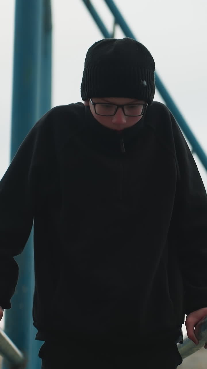 Close-up of a young boy finishing a workout on a metal bar, dressed in a black outfit and glasses, after dropping from the bar, he walks away