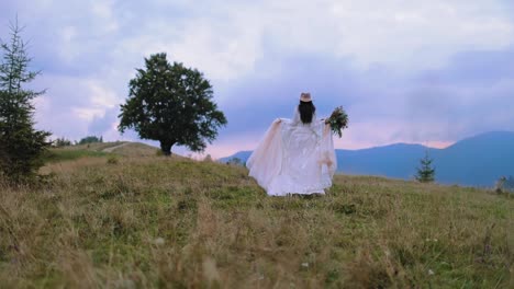 backside view of a bride in the mountains. woman in luxury wedding dress and hat walking among beautiful summer landscape.