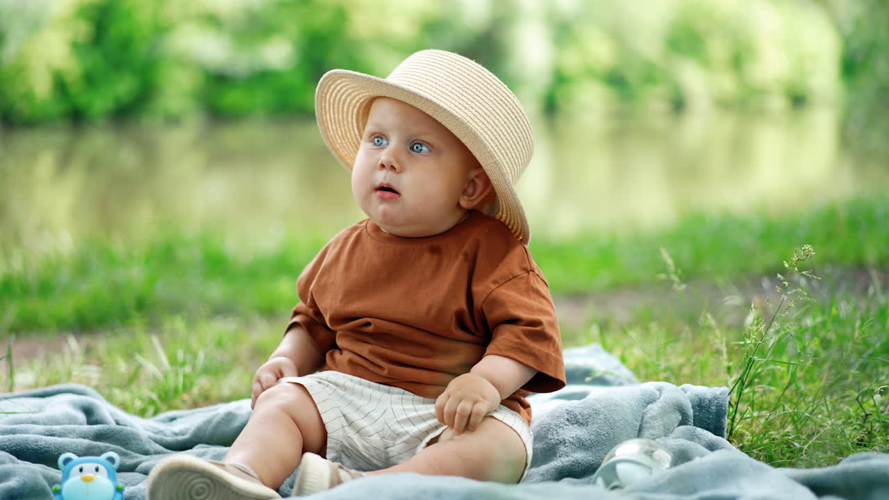 Beautiful baby boy with grey eyes wearing big straw hat sits outdoors. Child on the plaid chews something and looks up. Blurred backdrop.