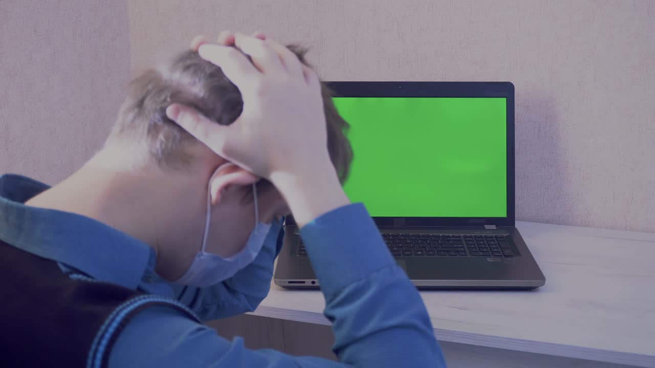 un tipo con una máscara protectora está emocionalmente enojado por una conexión remota en una videoconferencia y golpea sus puños en una computadora portátil, pantalla verde