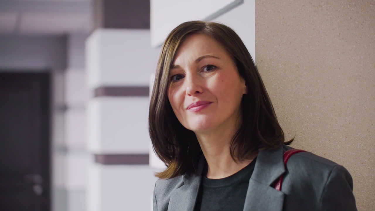 Confident young woman in business attire smiling at camera in modern office environment, exuding professionalism, standing by wall with stylish interior decor and natural light