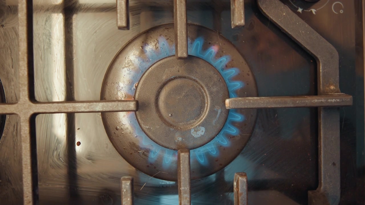 Primo piano dell'accensione di un fornello a gas per cucinare, la griglia metallica sporca si accende in una fiamma blu costante, espansione graduale della fiamma attorno al bruciatore circolare, la superficie del piano cottura mostra fuliggine e usura, luce calda della cucina, ideale.