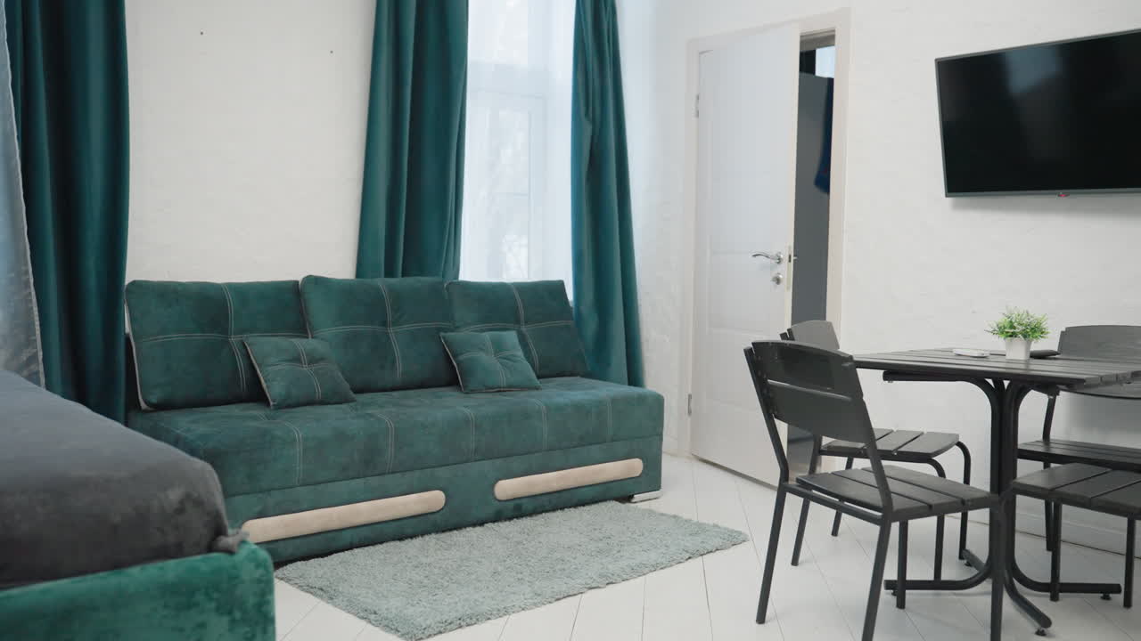 Modern apartment with green velvet sofa, black dining table, TV mounted on white wall, and small potted plant. Kitchen area visible in the background, creating a cozy and stylish living space
