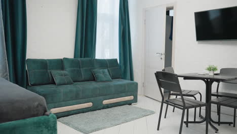 modern apartment with green velvet sofa, black dining table, tv mounted on white wall, and small potted plant. kitchen area visible in the background, creating a cozy and stylish living space