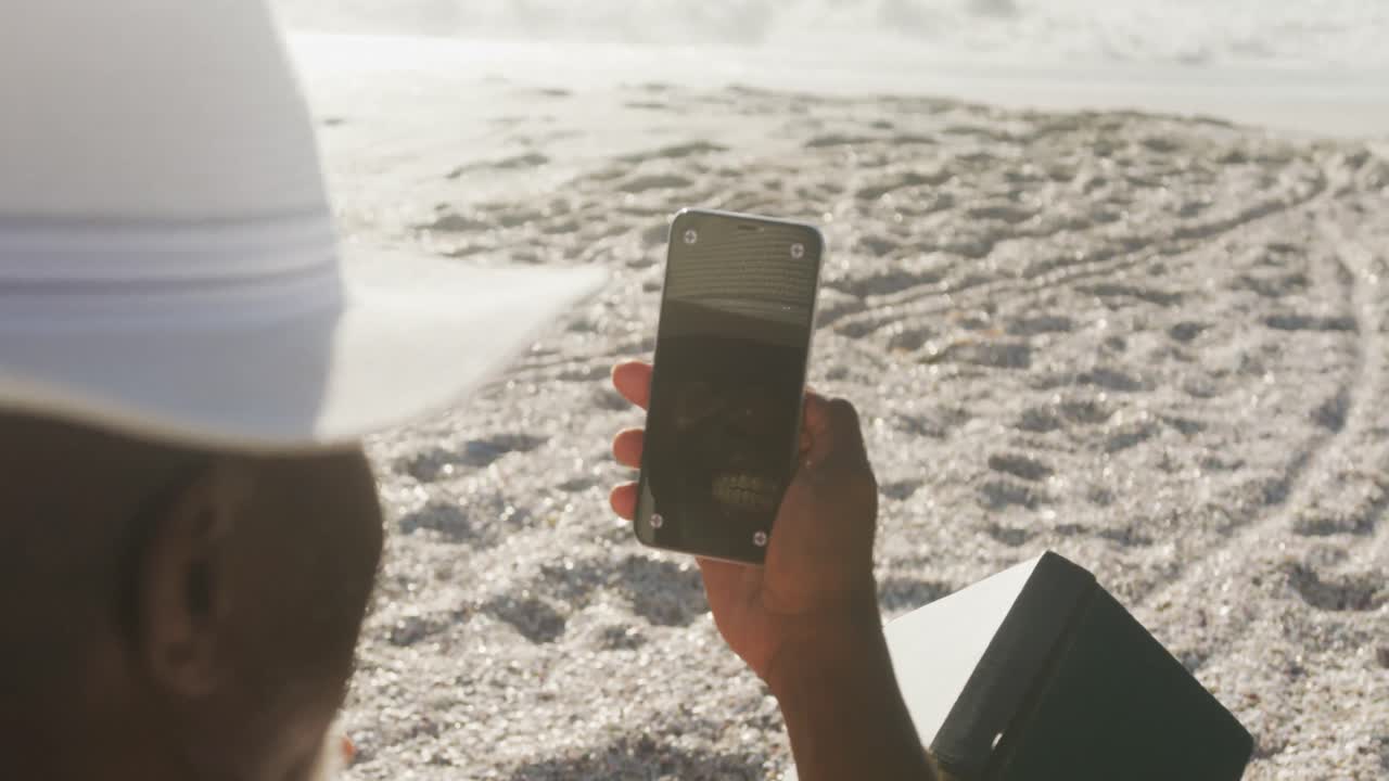 시니어 아프리카계 미국인 남자가  ⁇ 자리에 누워서  ⁇ 은 해변에서 스마트폰을 사용하고 있습니다.