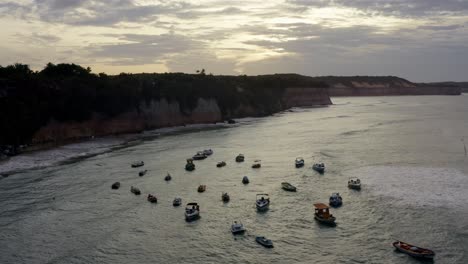 Un-Dron-Aéreo-En-Ascenso-Sobrevoló-Un-Grupo-De-Pequeños-Barcos-De-Pesca-Atracados-En-La-Playa-Tropical-De-Pipa-Durante-La-Hora-Dorada-Rodeada-De-Grandes-Acantilados-En-Río-Grande-Do-Norte,-Brasil