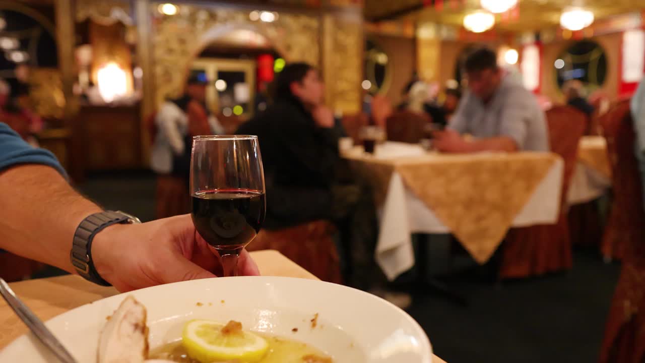 A person’s hand reaches for a glass of red wine on a table with a finished meal, set in a warmly lit, upscale restaurant with blurred diners in the background