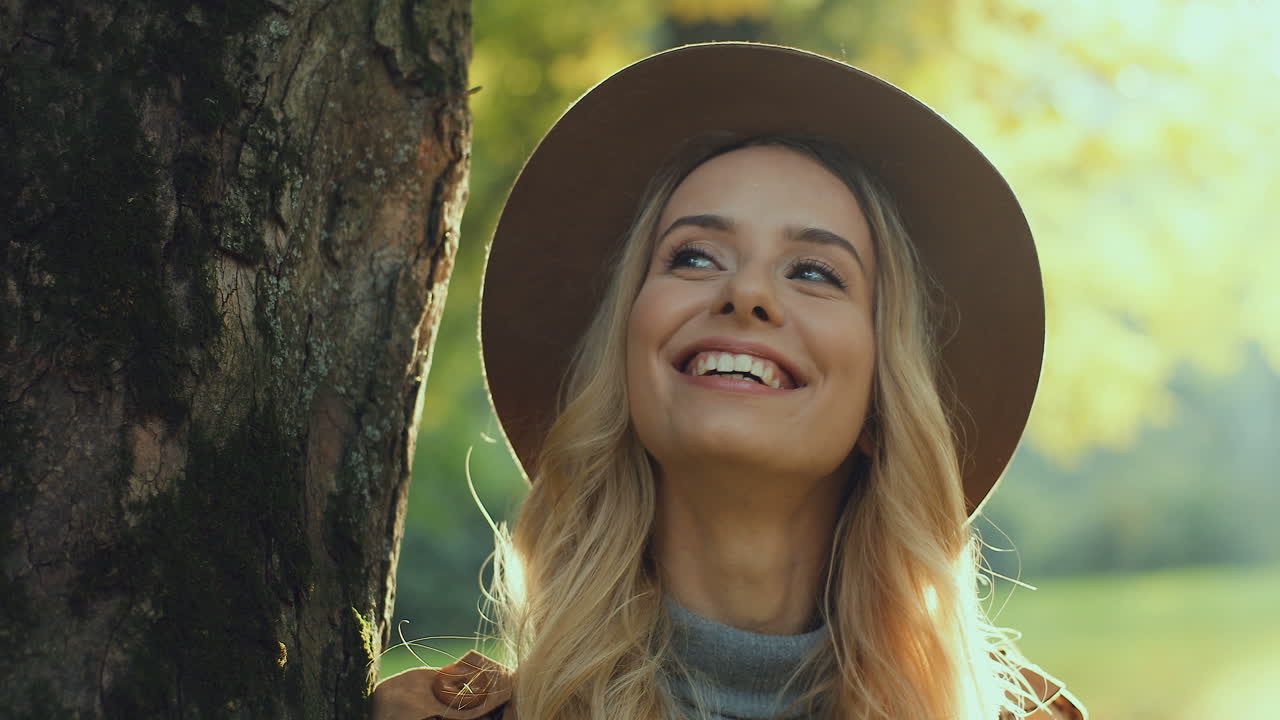 vista de cerca de una mujer rubia con sombrero, apoyada en un árbol y mirando alrededor con una sonrisa en la cara en el parque en otoño