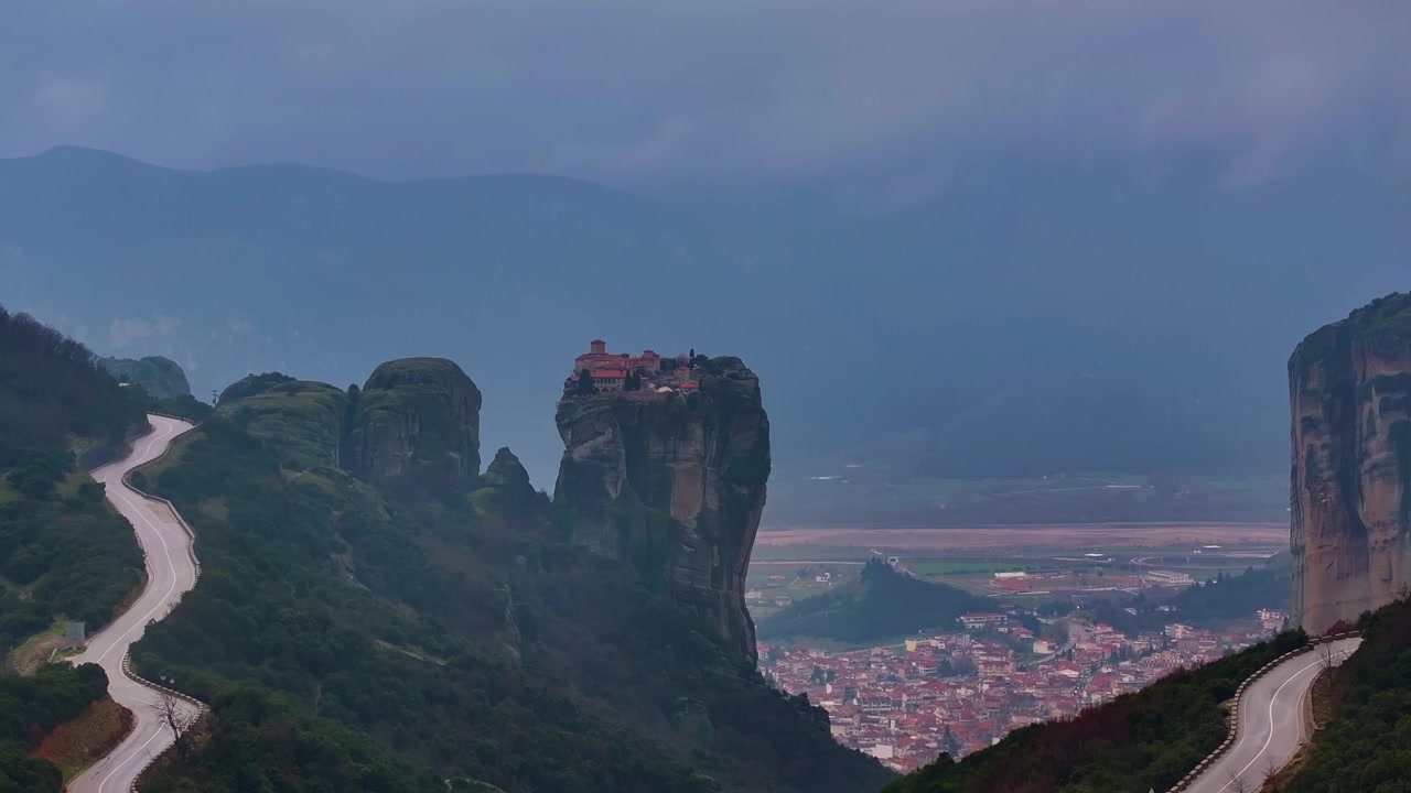 Bredt teleobjektivbillede af Hellig Treenigheds Kloster på toppen af en klippesøjle med snoede bjergveje og byen Kalambaka i dalen under stemningsfuld himmel i Meteora, Thessalien, Grækenland