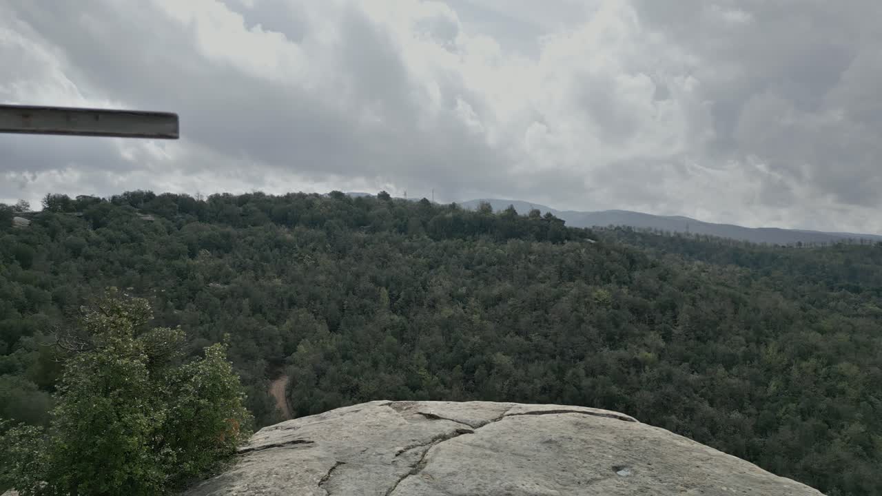 Metal cross standing on rocky mountain peak, overlooking vast lush green forest beneath cloudy sky, revealing expansive wilderness landscape with serene natural beauty