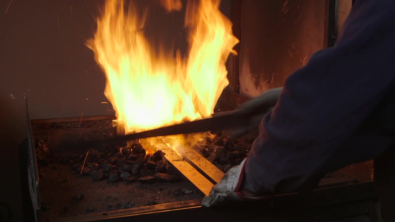 Close-up of a skilled blacksmith placing a steel bar into a traditional coal forge as intense flames surround the hot metal during the forging process.