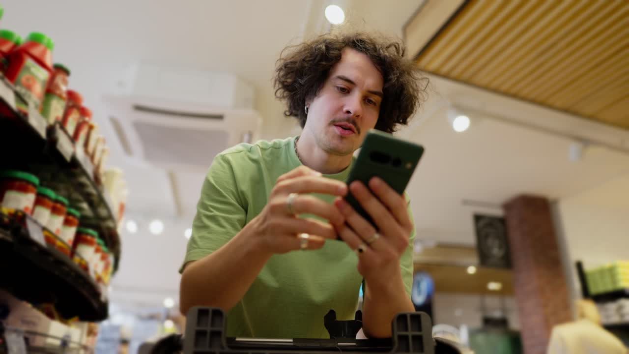 Confident guy with curly brunette hair wearing a green T-shirt scrolling through social networks while shopping in a supermarket with a cart