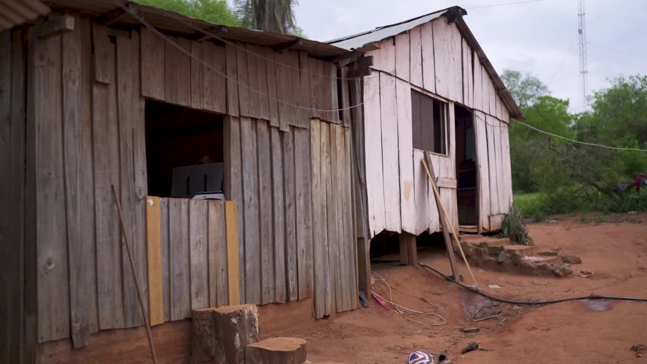 Two wooden structures stand side by side in a rural Mbya Guaraní community in Misiones, Argentina, built from vertical planks and elevated above red earth, with loose electrical wires and simple steps