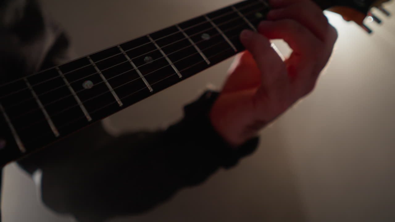 Close-up of a guitarist's hand strumming the strings of a guitar, highlighted by focused light in a shadowy room
