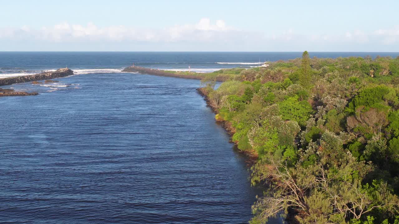 Drone footage captures a serene river flanked by lush greenery, leading to the ocean under clear skies