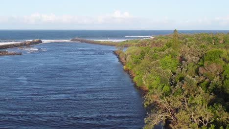 drone footage captures a serene river flanked by lush greenery, leading to the ocean under clear skies