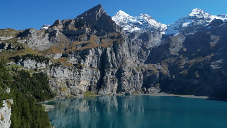 Fantastica-Toma-Aerea-Sobre-El-Lago-Oeschinen-Y-Pasando-Entre-Los-Arboles-Del-Bosque-Y-Las-Montañas-Que-Lo-Rodean,-En-Un-Dia-Soleado-Y-Apreciando-Las-Aguas-Turquesas