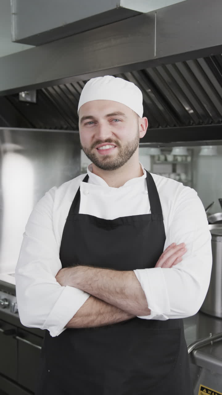 Happy caucasian male chef standing with arms crossed in kitchen, slow motion, vertical