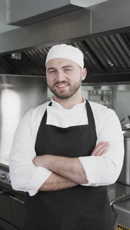 happy caucasian male chef standing with arms crossed in kitchen, slow motion, vertical