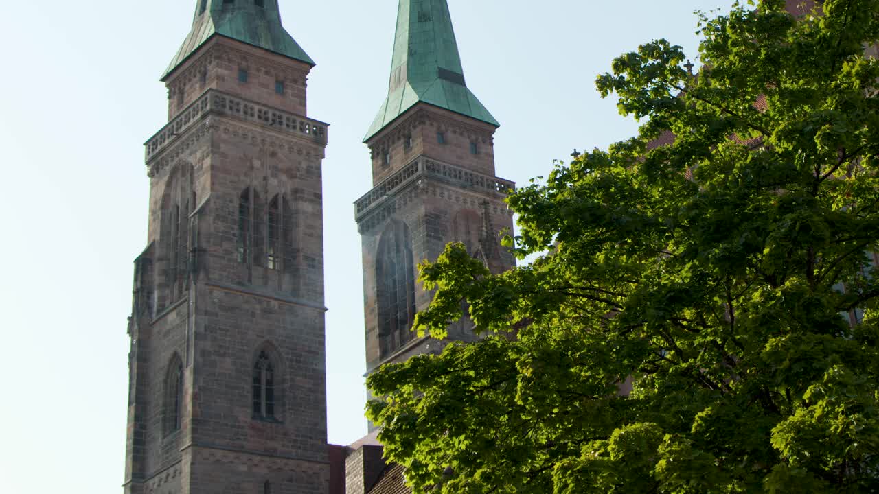 Camera tilts upward revealing historic church spires above green trees in bright daylight