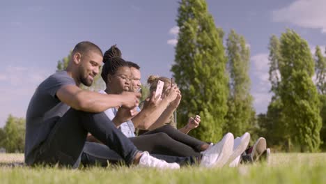 Happy-friends-sitting-on-green-lawn-and-taking-self-portraits