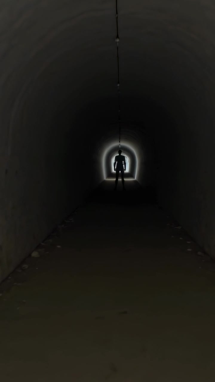 Video verticale: Luce che illumina una figura di fronte in abiti scuri che avanza in un tunnel con un cavo.
