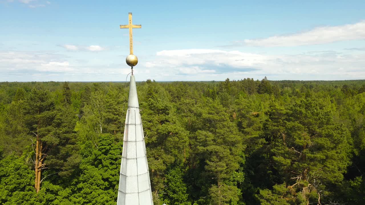 Aerial drone footage orbiting and spinning around a golden shiny and old cross on top of a church tower in the middle of a thick green forest with treetops and horizon visible in the background, sunny
