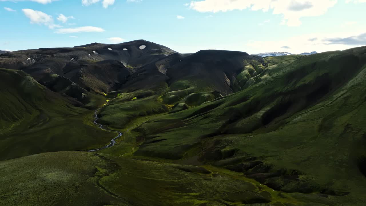 Aerial drift through a moss-covered valley, tracing a winding stream as it cuts between dark volcanic ridges under soft morning light in Iceland’s remote highland wilderness