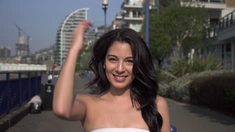 Slow-Motion-of-an-attractive-and-playful-latina-woman-with-black-wavy-hair-walking-on-the-banks-of-the-Thames-river-in-London,-looking-at-the-camera,-spinning,-happy-with-a-beautiful-smile