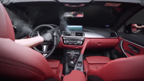 mecánico con un guante de goma negro y sosteniendo una aspiradora de vapor caliente limpiando las rejillas de ventilación del sistema de aire acondicionado en un lujoso interior de coche de cuero rojo