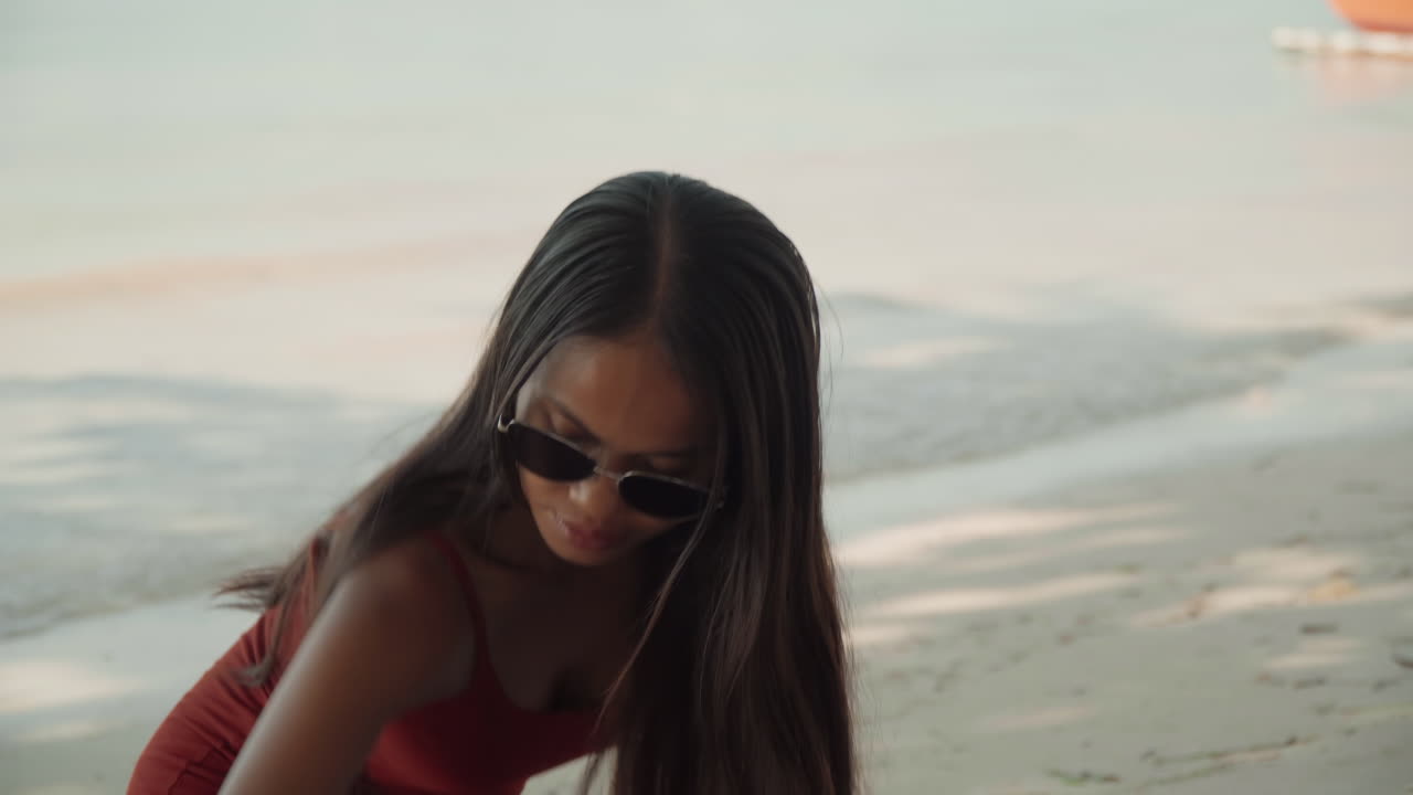 Giovane donna in spiaggia che indossa occhiali da sole, lunghi capelli intrecciati e prendisole rosso, onde leggere che lambiscono la riva, luce solare delicata, sorriso contemplativo, che si prepara per un'intima cerimonia del tè in riva al mare, spontanea.